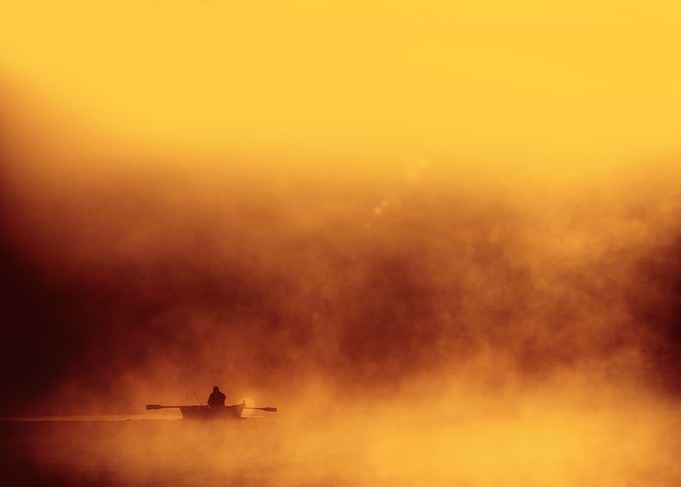 Lone fisher boat floating in foggy morning warm red yel ...