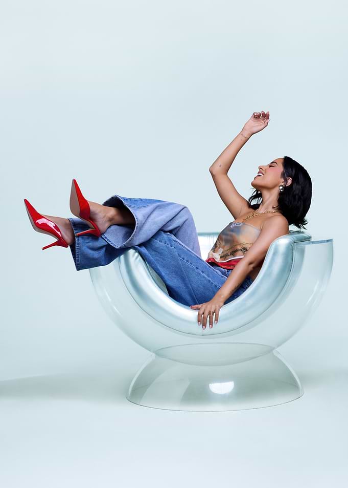 Becky G in jeans and heels in a clear chair