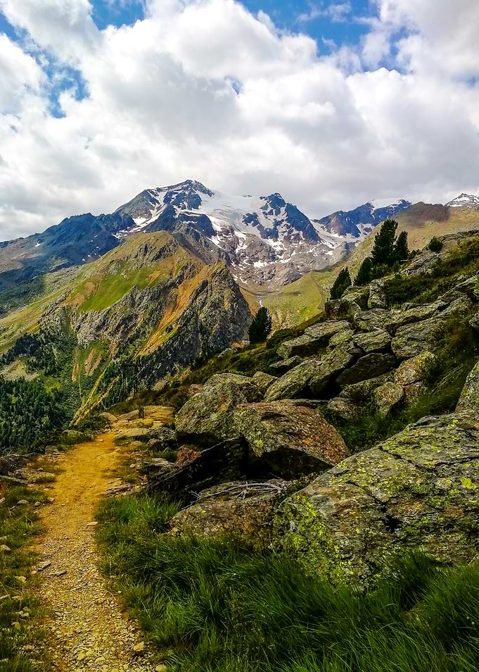 Narrow Mountain Trail Along a Rocky Ridge, with Snow-Capped Peaks and Dramatic Clouds, in the Italian Alps