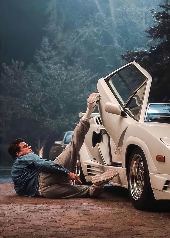 The image depicts Leonardo DiCaprio lying on the ground next to a white Lamborghini Countach with the door open. He is wearing a blue denim shirt, grey sweatpants, and white sneakers. 