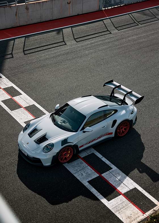 A white Porsche 911 GT3 RS, featuring a large rear wing and vibrant orange-red wheels, is positioned on a dark asphalt race track. 