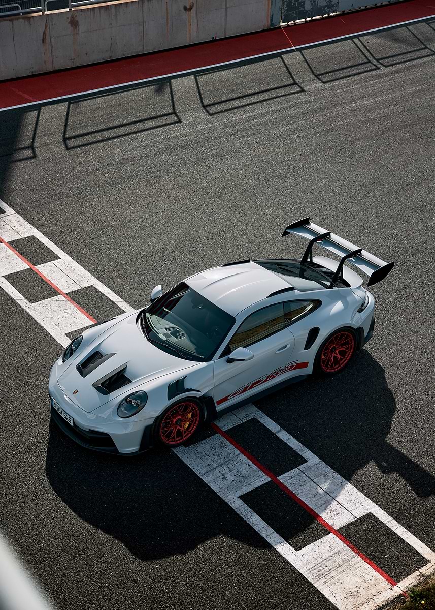 A white Porsche 911 GT3 RS, featuring a large rear wing and vibrant orange-red wheels, is positioned on a dark asphalt race track. 